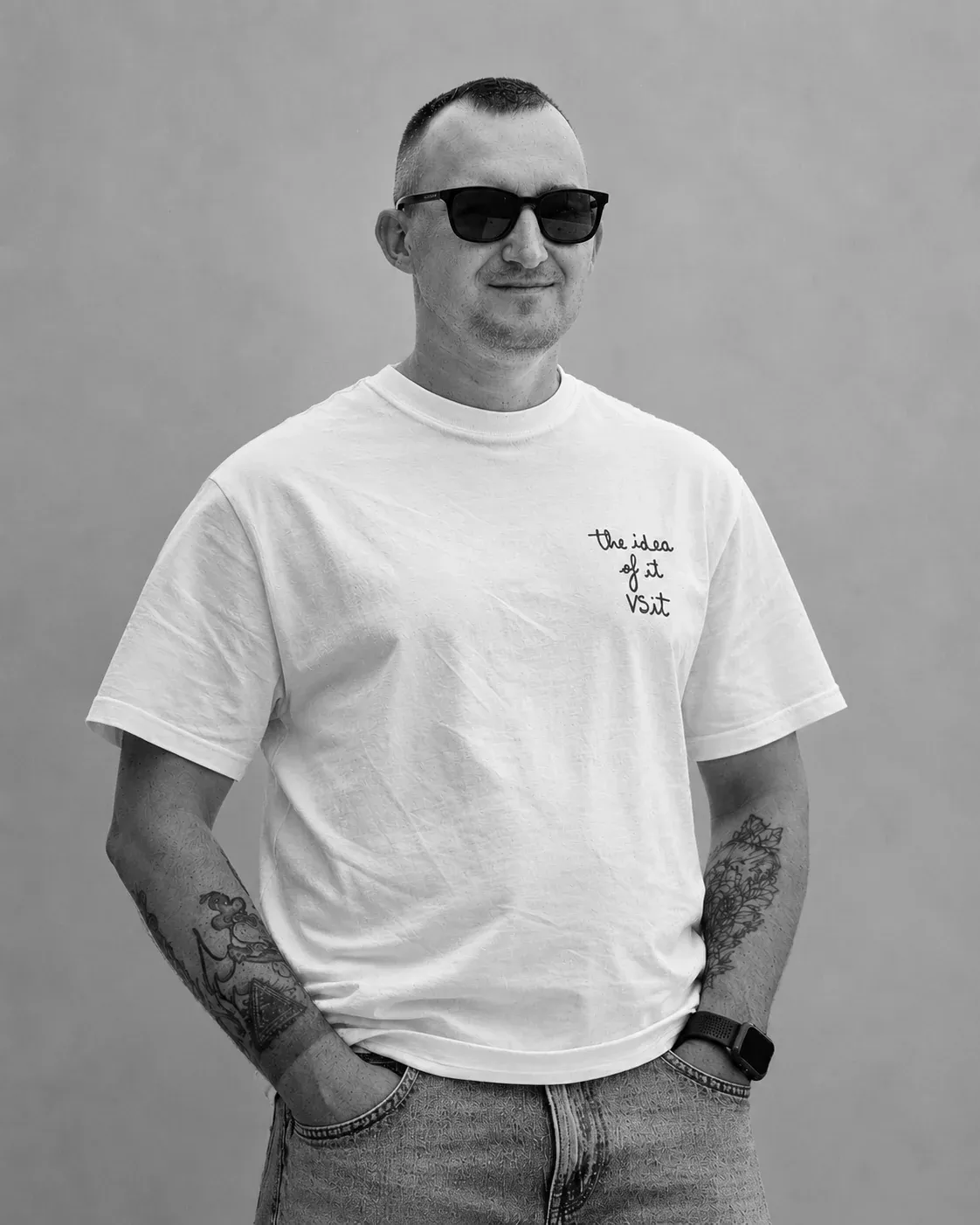 Max Chechel in sunglasses and a white T-shirt, hands in his pockets, against a grey wall.